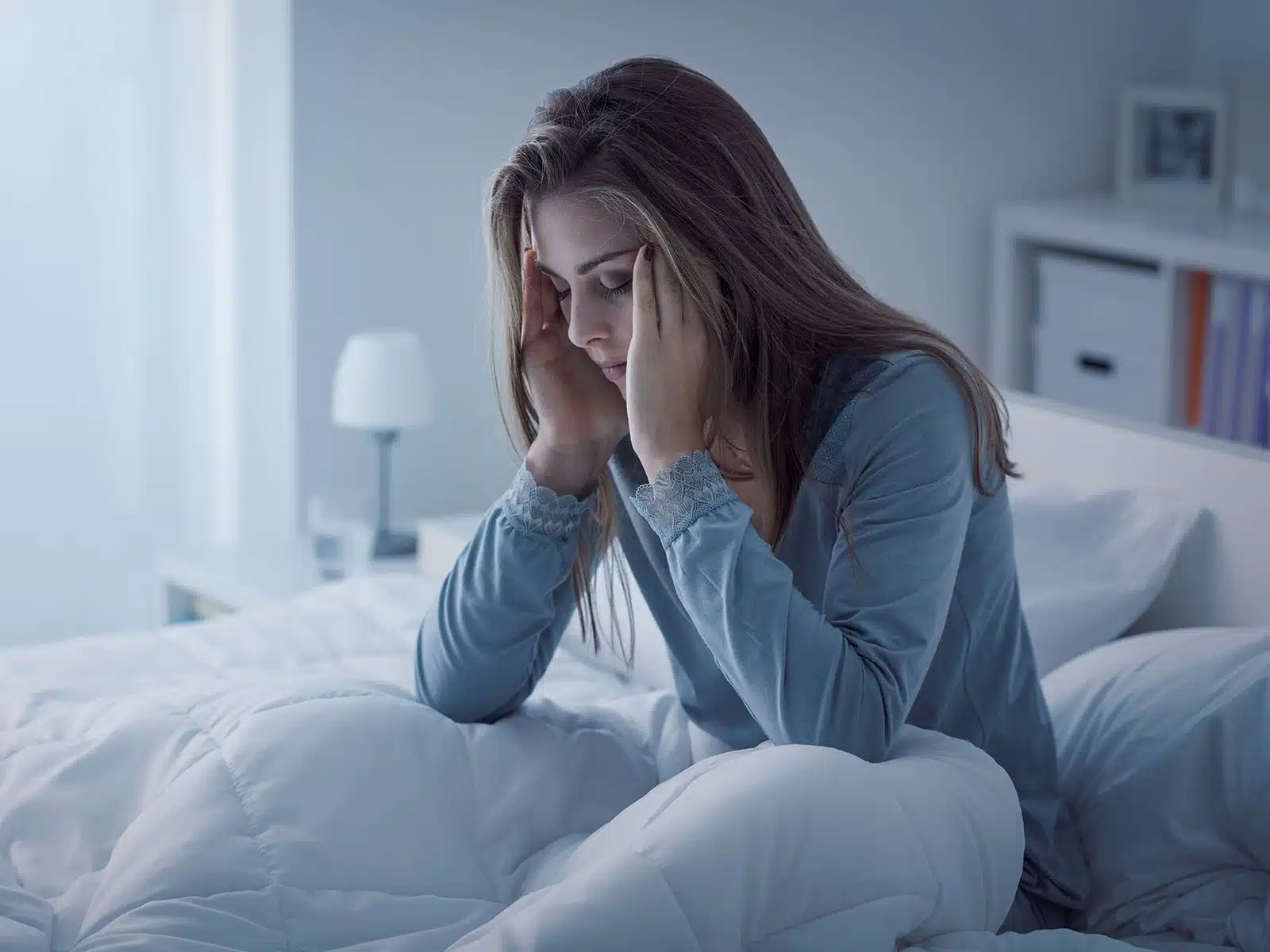 A woman sits up in bed at night, holding her head with both hands, looking distressed or unable to sleep.