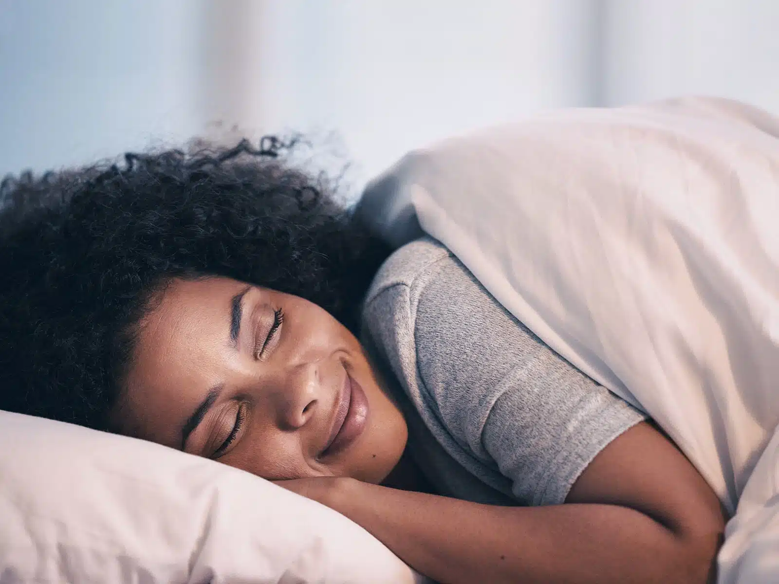 A woman with curly hair sleeps peacefully, smiling slightly, wrapped in a blanket.