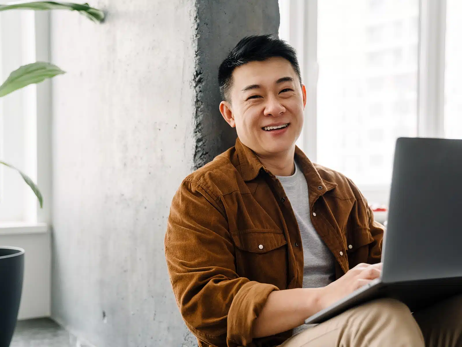 A smiling man in a brown jacket sits with a laptop, leaning against a concrete wall in a bright room.