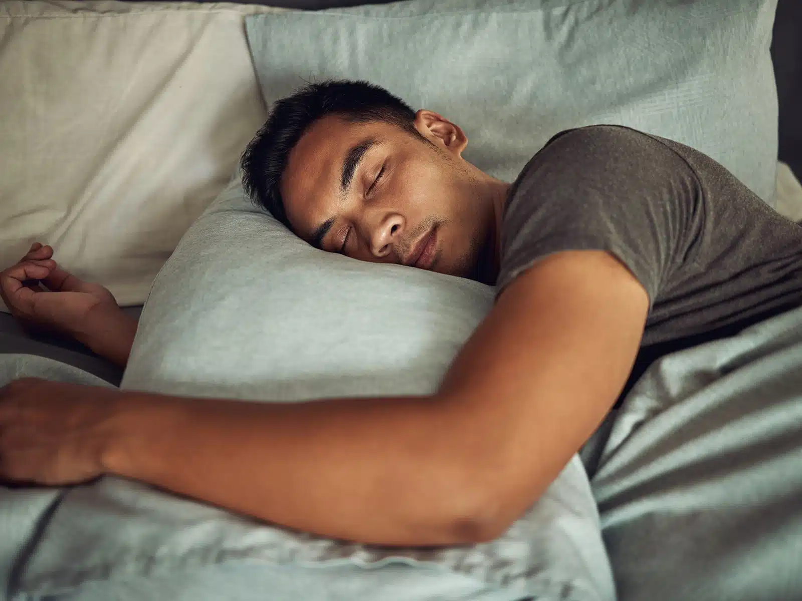 A young man in a dark t-shirt sleeps peacefully on his side, resting his head on a light blue pillow.