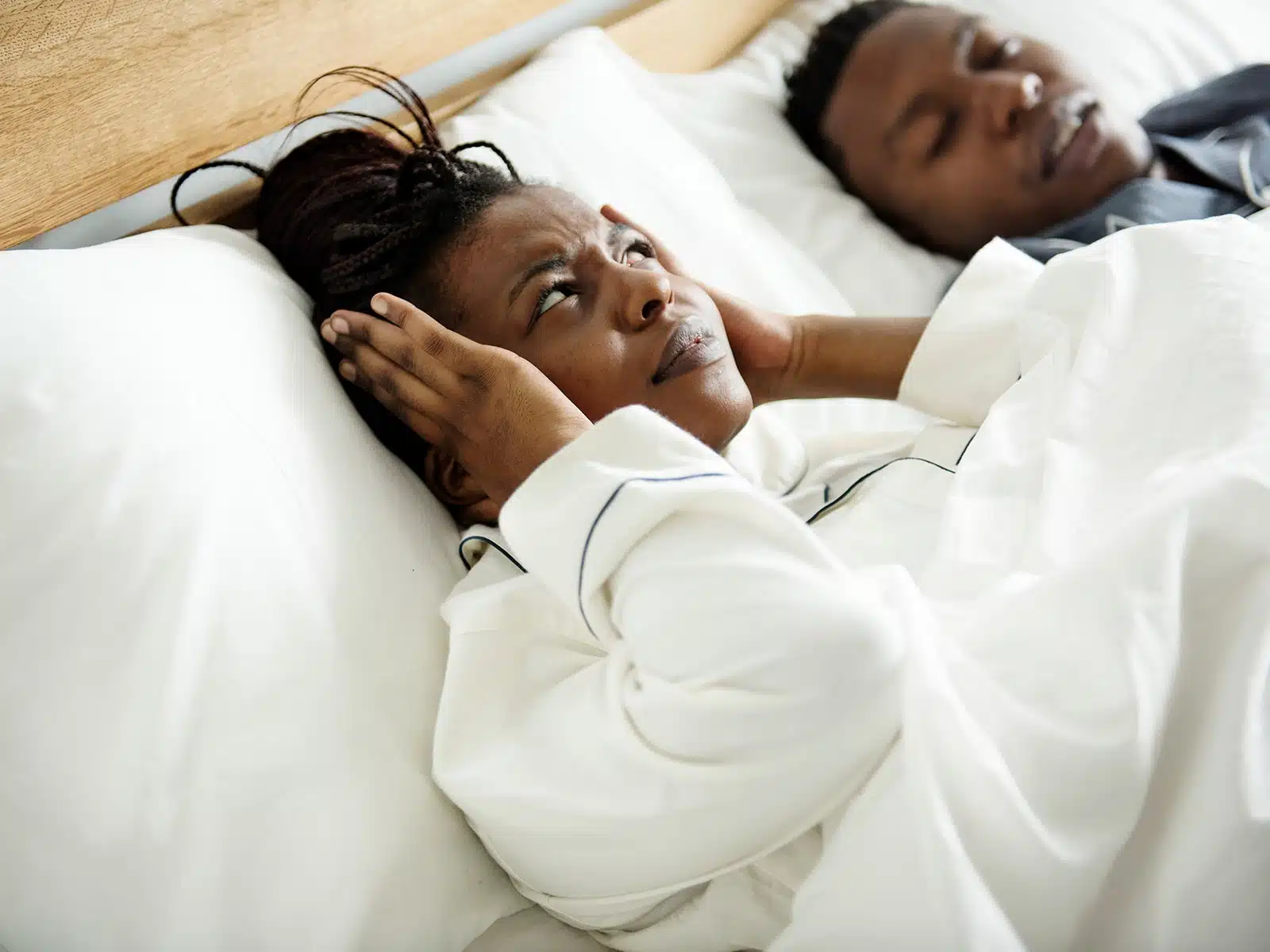 A woman in bed covers her ears with a frustrated expression while a man beside her sleeps peacefully, possibly snoring.