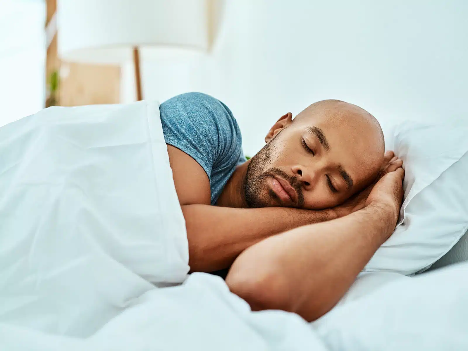A man in a blue shirt sleeps peacefully on his side, resting his head on his hands under a white blanket.