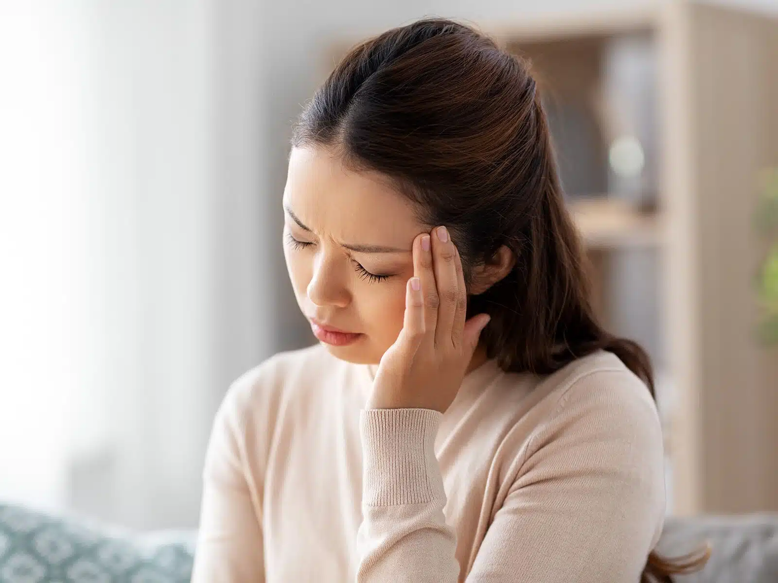 A woman sits indoors, pressing her temple with a pained expression, appearing to have a headache or discomfort.