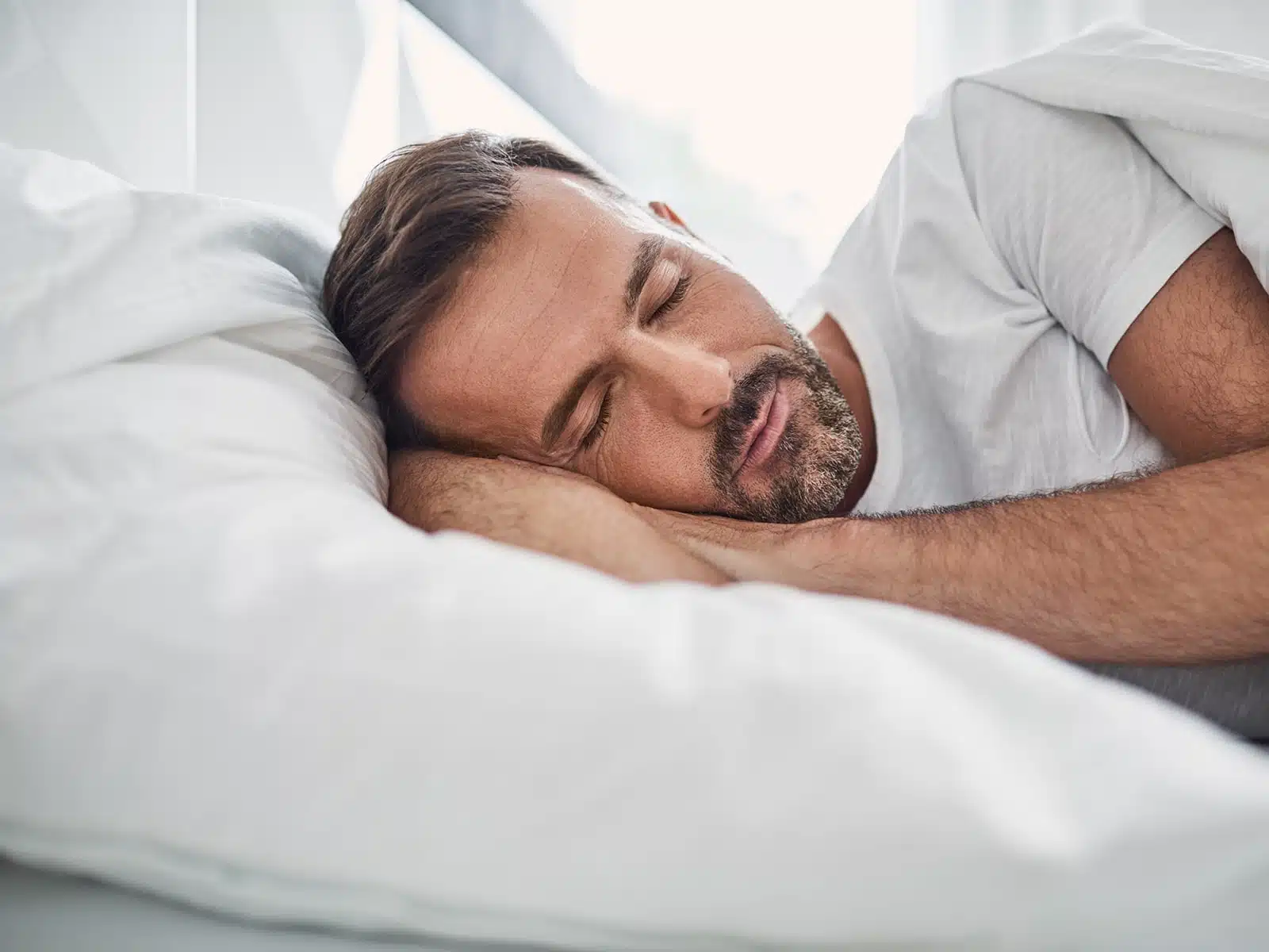 A man with a beard sleeps peacefully on his side, resting his head on a white pillow with a relaxed expression.