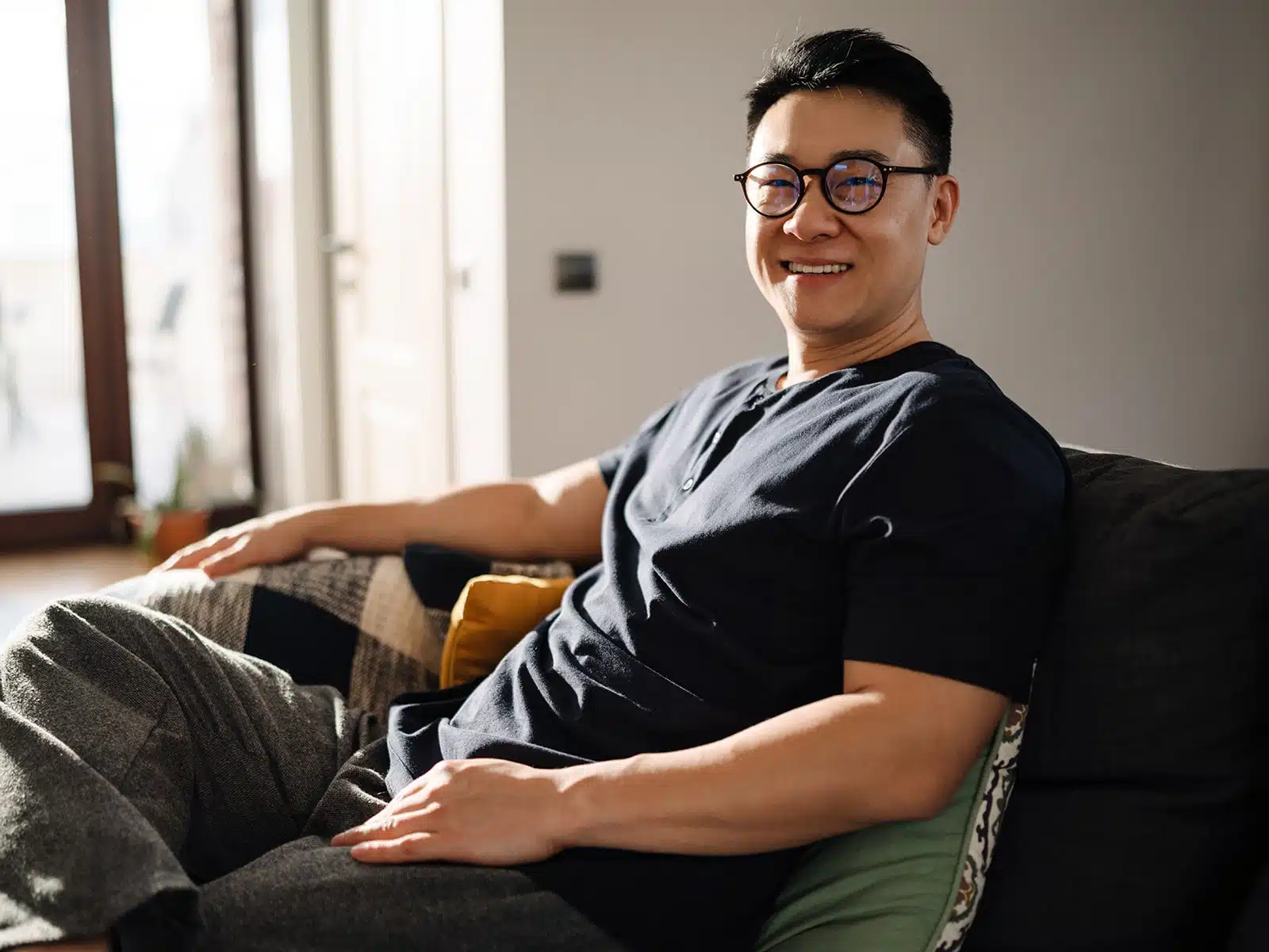 A man wearing glasses and a black shirt sits on a couch, smiling warmly while enjoying the sunlight streaming in from a nearby window.