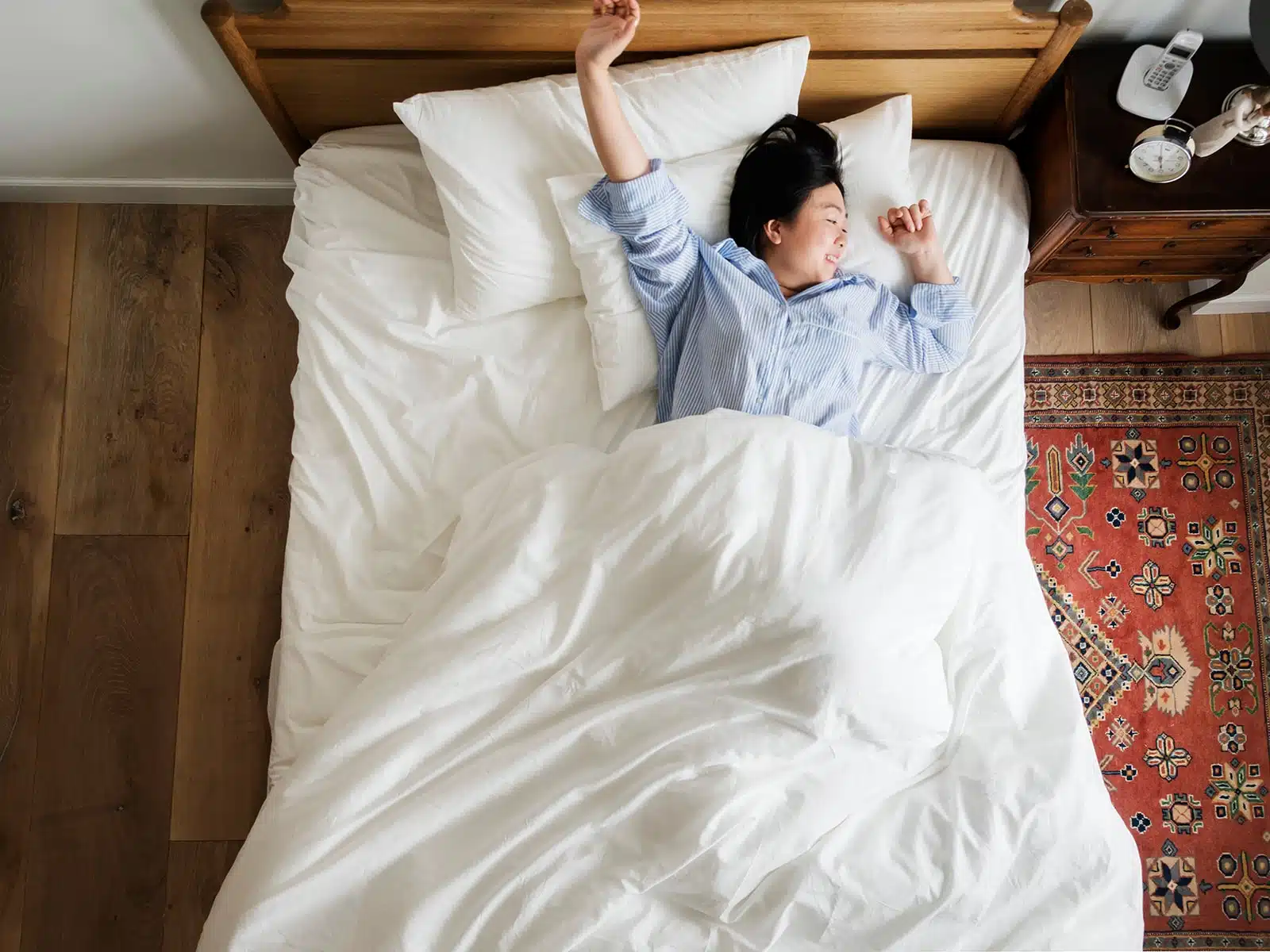 A woman in a blue striped pajama shirt stretches and smiles while waking up in a cozy bed with white bedding.