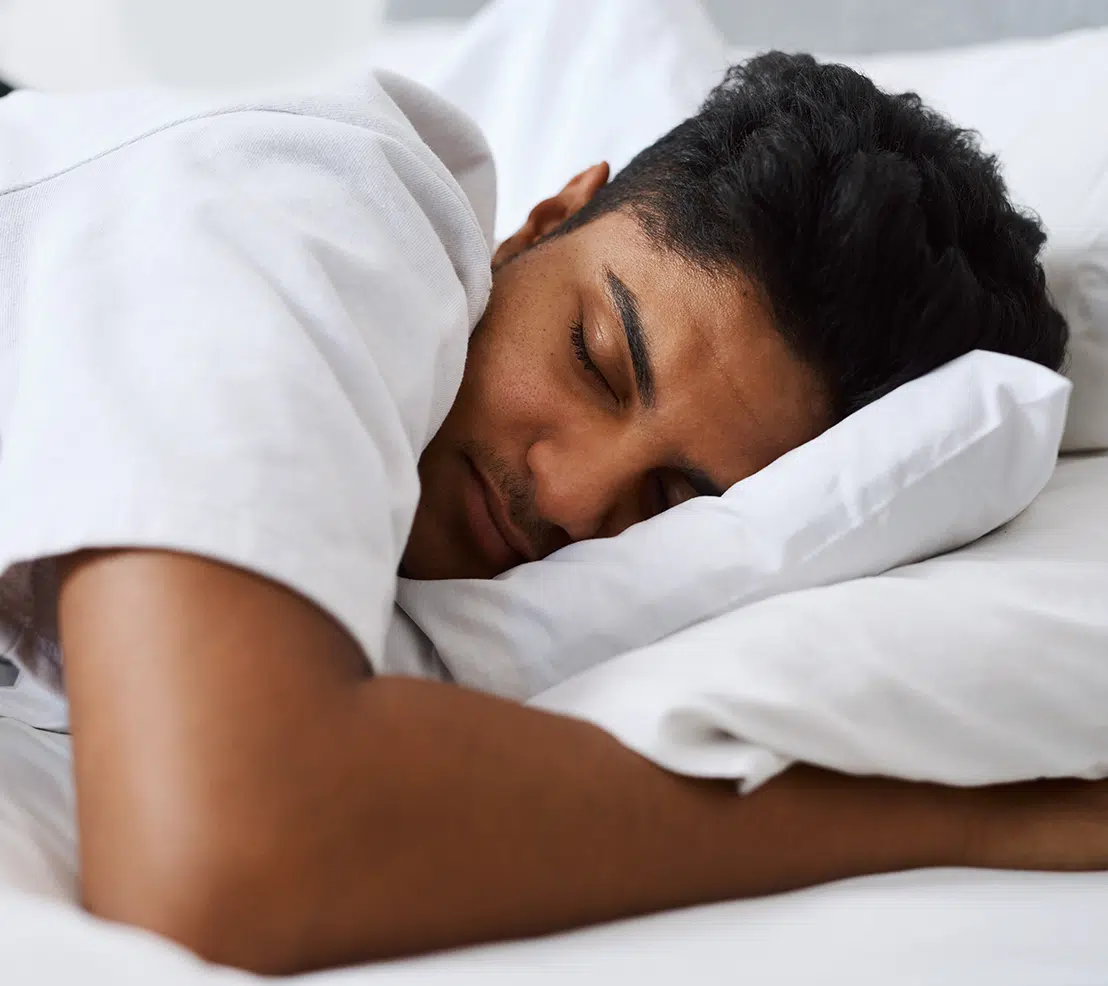 A man in a white shirt sleeps peacefully on his side, resting his head on a white pillow.