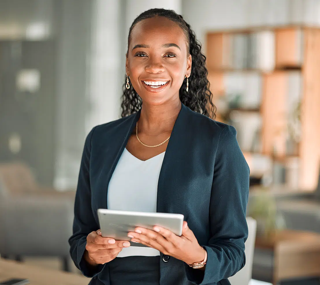 A confident woman in a navy blazer smiles while holding a tablet in a modern office setting.