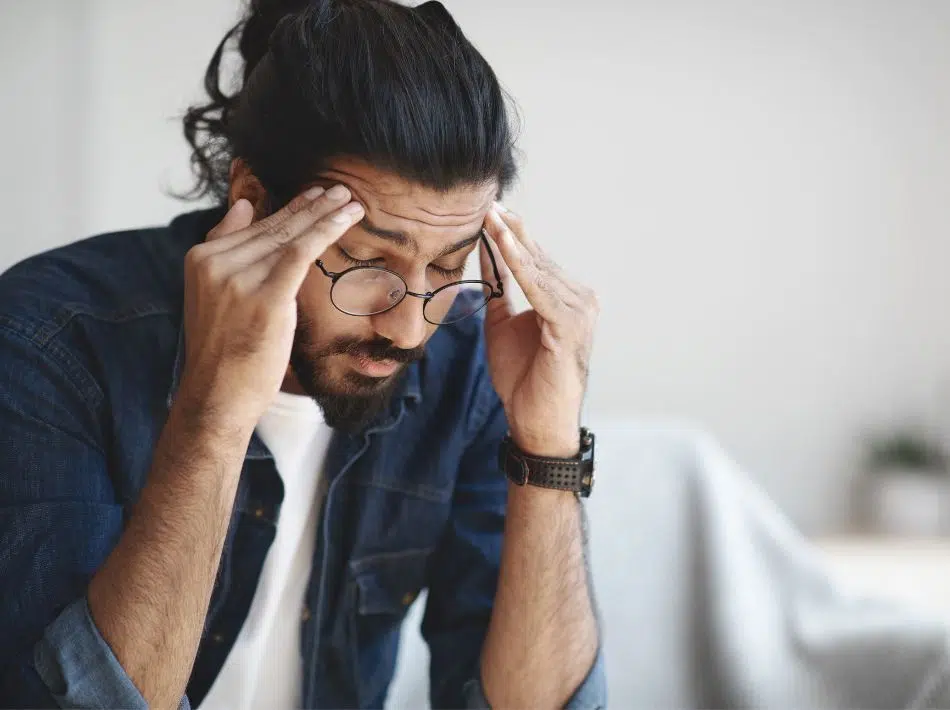 A man with long, dark hair and glasses holding his temples, clearly experiencing a headache